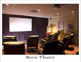 A small movie theater room with six black leather recliner chairs arranged in two rows facing a large white projection screen. The room has dim lighting, carpeted floor with a patterned design, and framed posters on the walls.