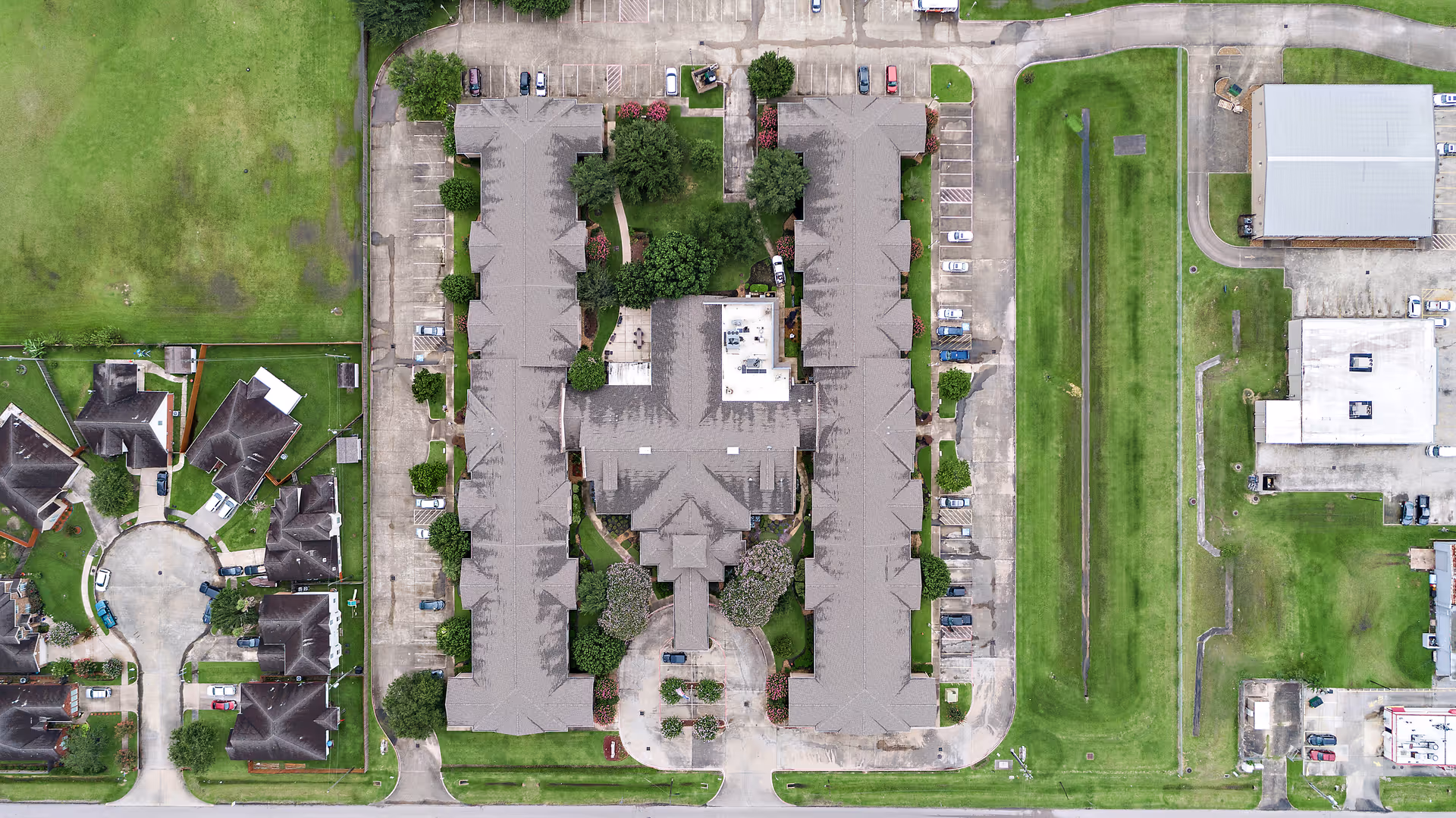 Aerial top-down view of a U-shaped residential complex with parking lots, trees, and surrounding lawns.