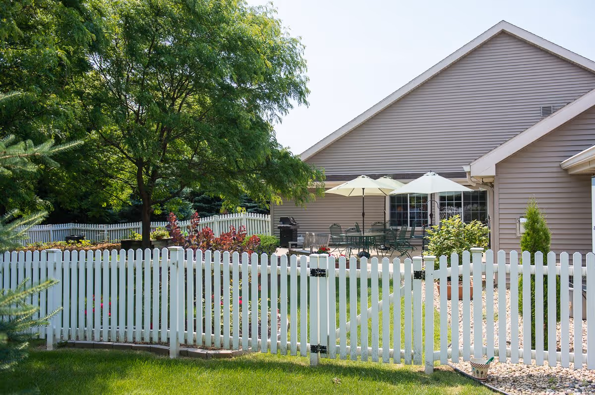 A fenced backyard area with a white picket fence, green grass, and a variety of plants and trees. There is a patio with several green metal chairs and tables under two large umbrellas next to a beige house with a sloped roof.