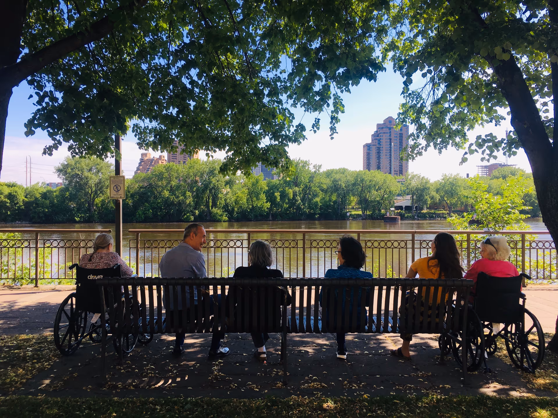 Six people sitting on a bench under the shade of trees by a riverside, with two individuals in wheelchairs on either end. Across the river, there are green trees and tall buildings in the background under a clear blue sky.
