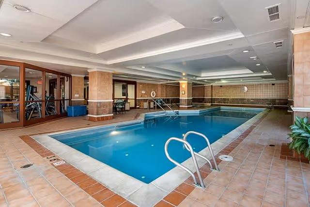 Indoor swimming pool area with tiled floors and walls, metal handrails for pool entry, and a seating area visible through glass doors on the left side.