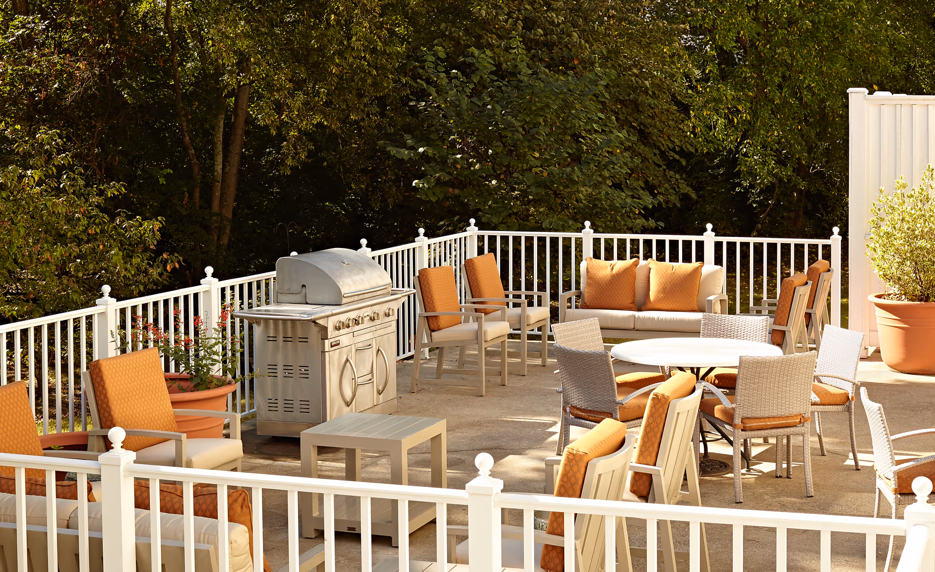 Outdoor patio area with a stainless steel grill, multiple cushioned chairs with orange cushions, a round table with chairs, a small square table, and a white railing surrounding the space. There are large potted plants and dense green trees in the background.