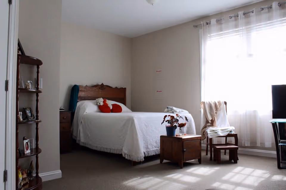 A bedroom with a wooden bed frame and white bedspread, featuring a red heart-shaped pillow and a white teddy bear. There is a wooden nightstand with a potted plant and a book, a wooden chair with a blanket draped over it, and a small wooden stool. Sheer white curtains cover a large window letting in natural light. A wooden shelf with framed photos and decorative items is visible on the left side.