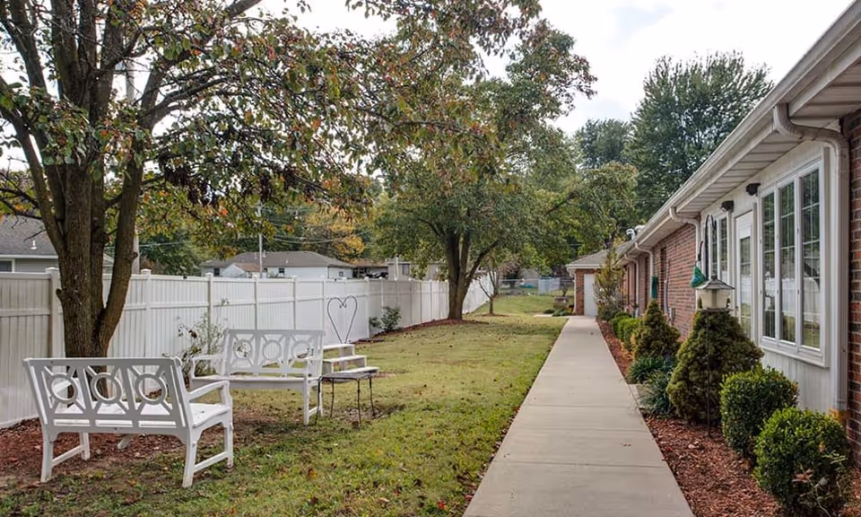 Outdoor view of a senior living facility with a concrete walkway alongside a brick building. There are two white benches and a small table under a tree on a grassy area next to a white fence. Shrubs and small trees line the building.