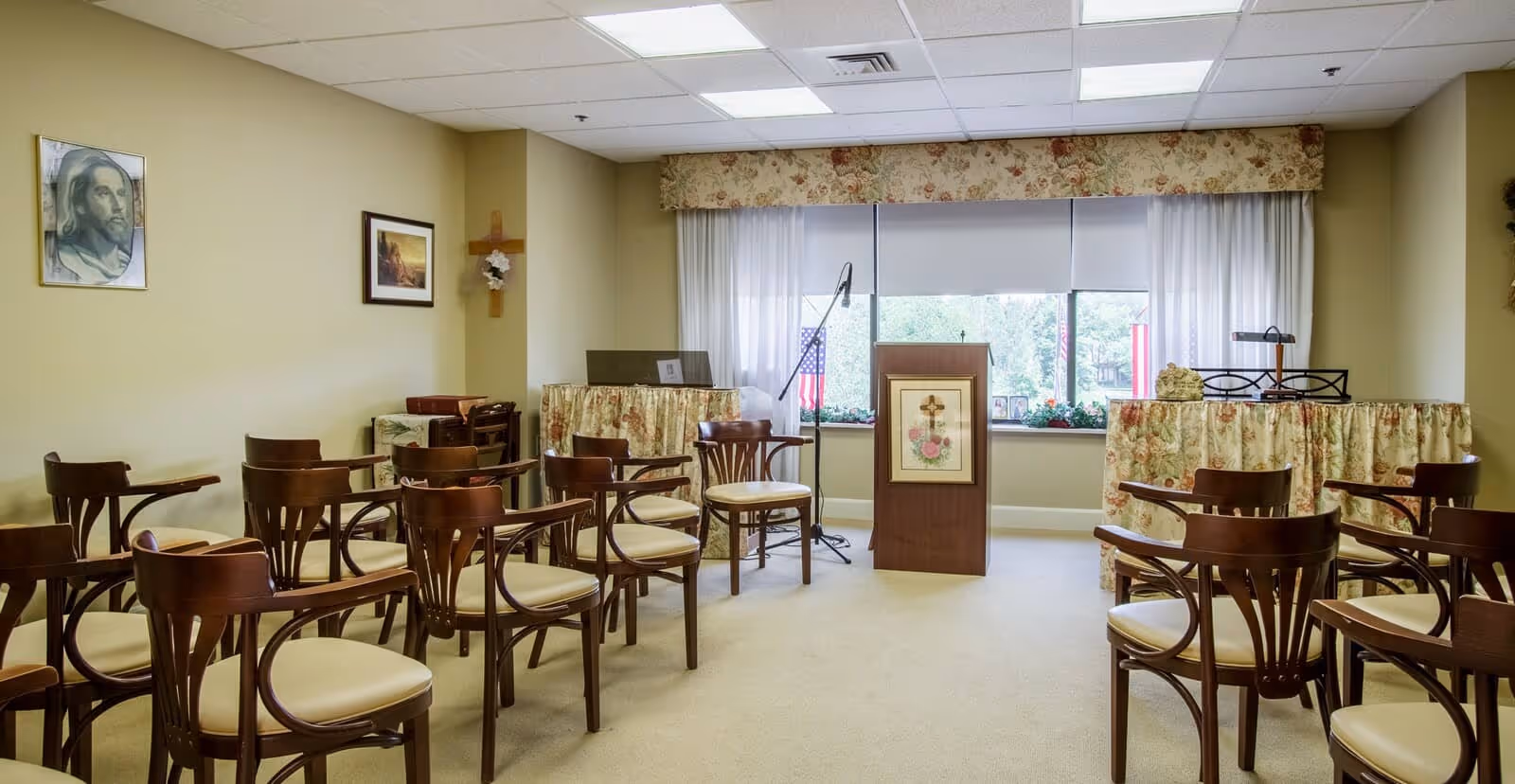 A small chapel or meeting room with rows of wooden chairs with beige cushions arranged facing a wooden podium decorated with a floral cross picture. Behind the podium is a window with floral valance and sheer curtains, and an American flag is visible outside. The room has beige walls adorned with religious artwork and a wooden cross with a flower decoration. There is a microphone stand next to the podium and tables covered with floral fabric on either side.