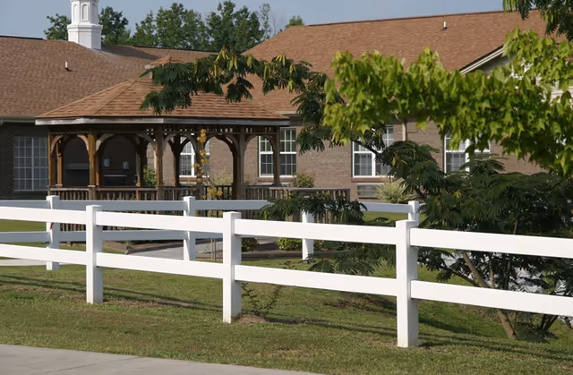 White fence and grassy lawn in front of a brick senior living building with a wooden gazebo and trees.