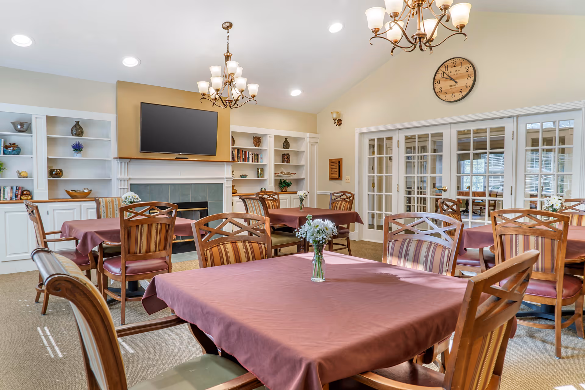 A dining room with several tables covered in purple tablecloths, each with a small vase of flowers. Wooden chairs with striped cushions surround the tables. The room features a fireplace with a mounted flat-screen TV above it, built-in white shelves with decorative items, a large wall clock, and glass-paneled double doors leading to another room.