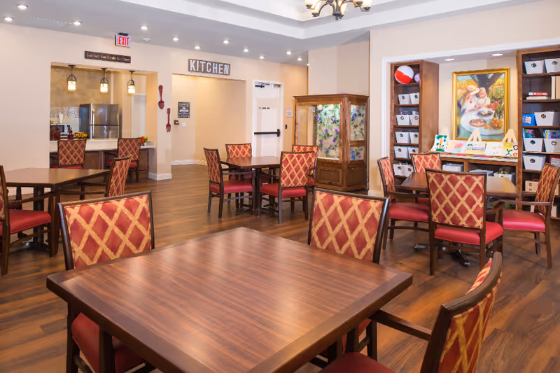 A dining area with multiple wooden tables and red cushioned chairs featuring a diamond pattern. The room has wooden flooring and beige walls. In the background, there is a kitchen area with stainless steel appliances visible through an open doorway marked with a 'KITCHEN' sign. Shelves with bins and a colorful painting are also visible on the right side of the room.