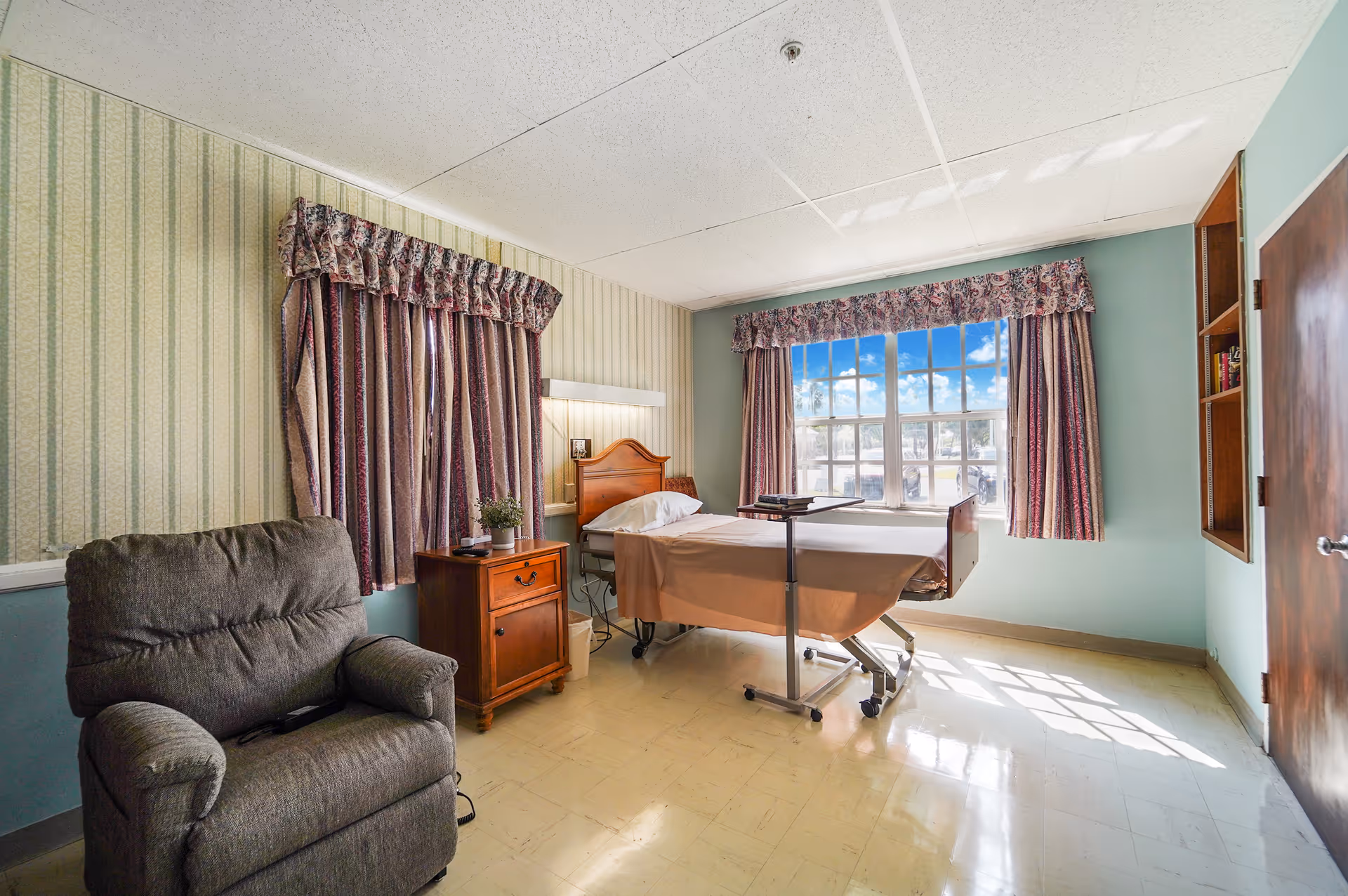 A well-lit room with a hospital-style bed covered with a beige sheet, positioned near a large window with floral curtains. Next to the bed is a wooden nightstand with a small plant and a remote control. A comfortable gray recliner chair is placed against the wall with striped wallpaper. The floor is light-colored linoleum, and there is a wooden door and built-in shelves on the right side of the room.