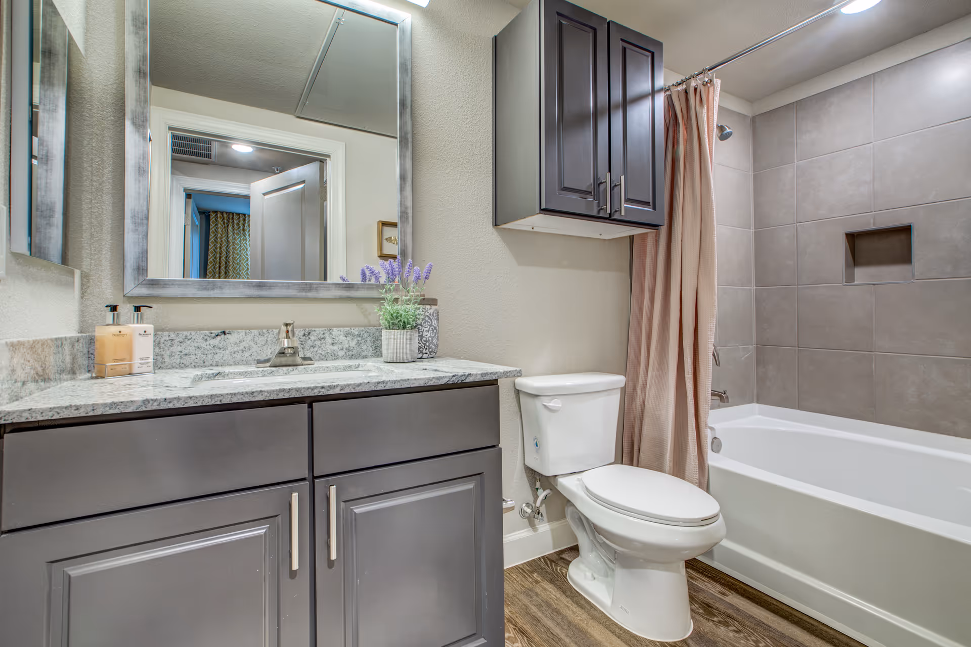 A clean and modern bathroom featuring a white toilet, a bathtub with a beige shower curtain, and a wall-mounted cabinet above the toilet. There is a granite countertop with a sink, a large rectangular mirror above it, and a small potted plant and soap dispensers on the counter. The floor has wood-like tiles and the walls are painted light beige.