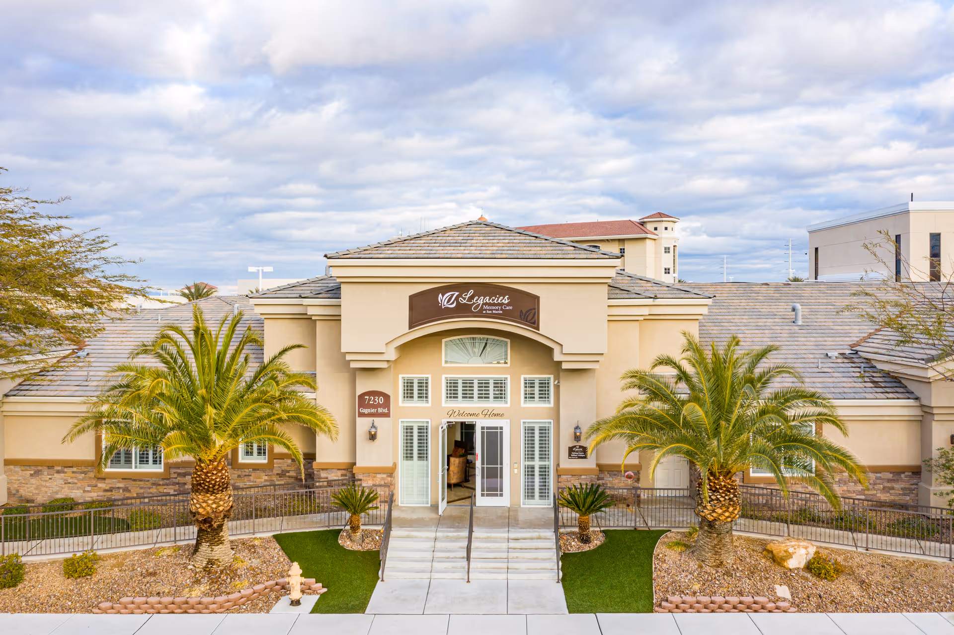 Front exterior view of Legacies Memory Care at San Martin building with a beige facade, two palm trees flanking the entrance, and a sign above the door. The entrance has glass doors and windows with white frames, and there is a ramp and stairs leading up to it. The sky is partly cloudy.