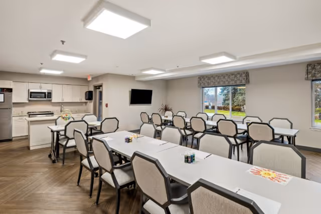 A bright, spacious room with multiple tables and chairs arranged in rows, set up for an activity with art supplies on the tables. The room has large windows showing an outdoor view, a wall-mounted TV, and a kitchenette area with white cabinets, a microwave, and a refrigerator.