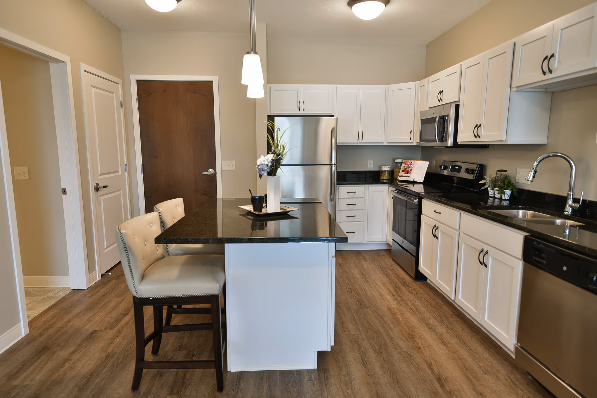 Modern kitchen with white cabinets, black granite countertops, stainless steel appliances including refrigerator, stove, microwave, and dishwasher. There is a kitchen island with two beige cushioned chairs and a vase with flowers on top. The floor is wood-style, and there are two pendant lights hanging above the island.