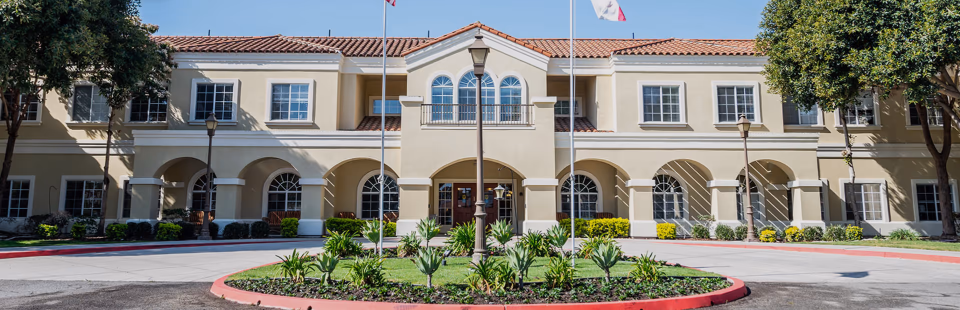Front exterior view of a two-story senior living facility building with beige walls, red tile roof, arched windows and doorways, a circular driveway with a landscaped island in the center, and two flagpoles in front.