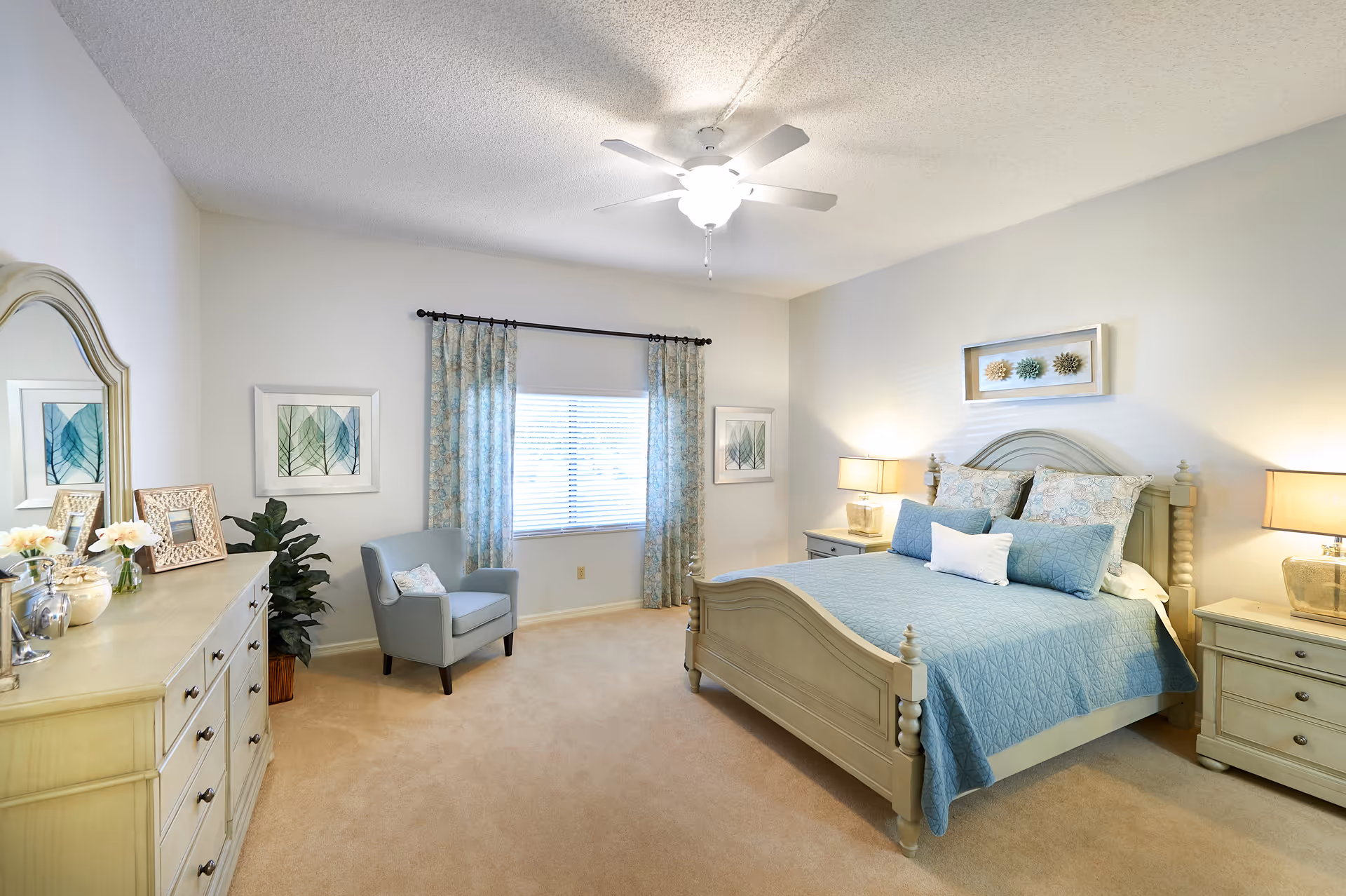 A bright and spacious bedroom with a large bed covered in blue bedding and multiple pillows. There are two matching nightstands with lamps on either side of the bed. A dresser with a mirror, decorative items, and picture frames is on the left side of the room. A comfortable armchair is positioned near a window with blue patterned curtains. The walls are light-colored, and the room has beige carpeting and a ceiling fan with a light.