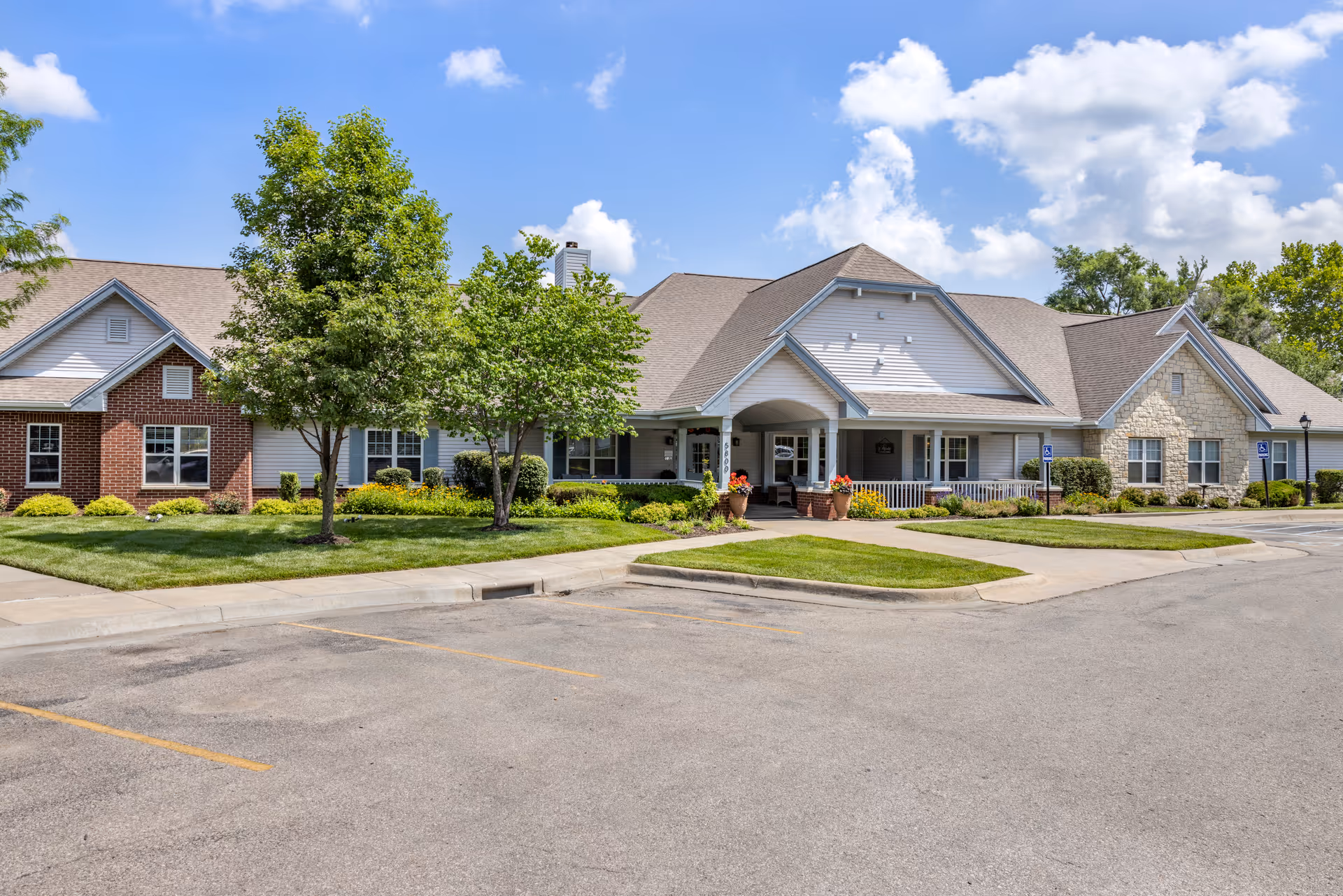 Exterior front view of a single-story senior living facility building with a combination of brick, stone, and siding. The building has a covered entrance with potted plants and a well-maintained lawn with trees and shrubs. The sky is blue with scattered clouds, and there is an empty parking lot in front.