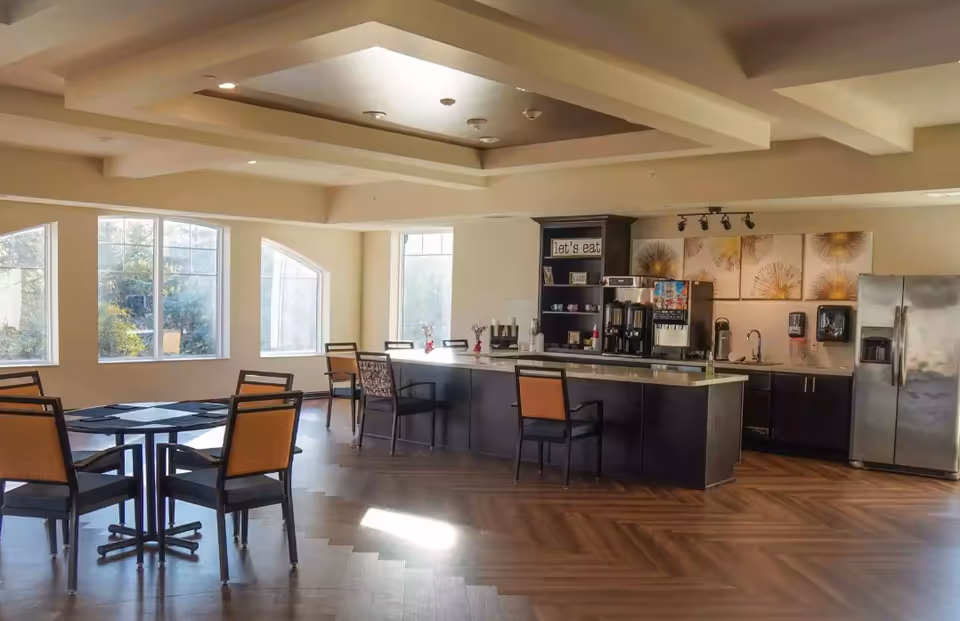 A bright dining area with large windows letting in natural light. The room features a few tables with chairs and a kitchen counter with stools. The kitchen area includes a refrigerator, coffee machines, and decorative wall art. The ceiling has a recessed design with lighting fixtures.