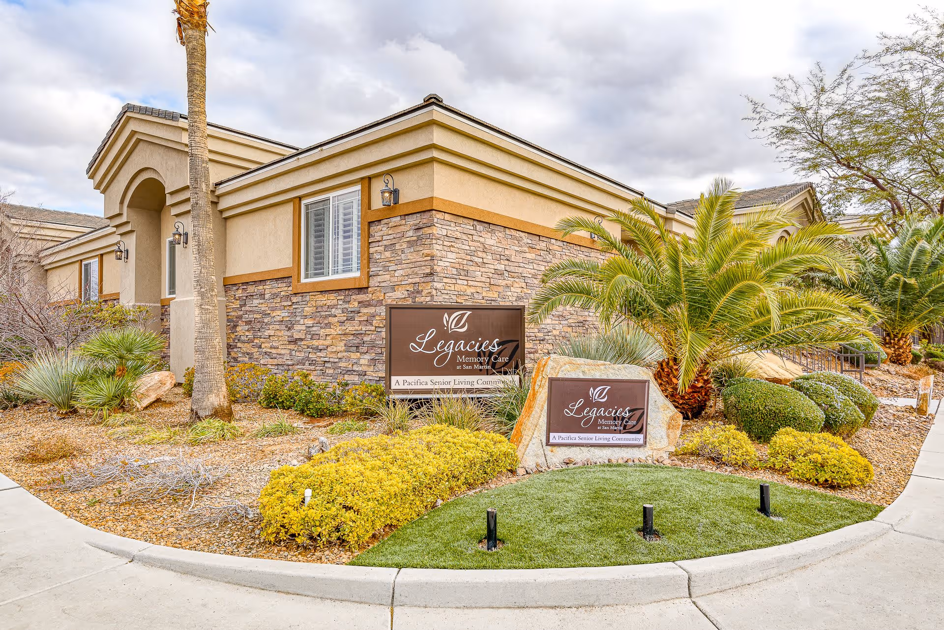 Exterior view of a senior living facility building with stone and beige stucco walls, surrounded by landscaped plants including palm trees and shrubs. Two signs in front read 'Legacies Memory Care at San Martin, A Pacifica Senior Living Community.'