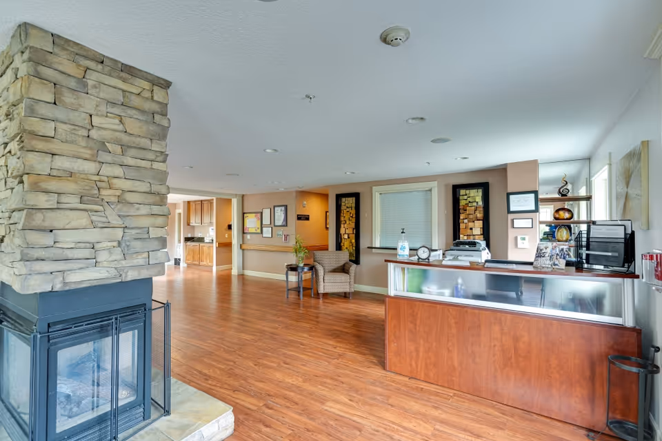 Interior view of a senior care facility reception area with a stone fireplace on the left, wooden flooring, a wooden reception desk on the right, a chair and small table with a plant in the middle, and framed artwork on the walls.