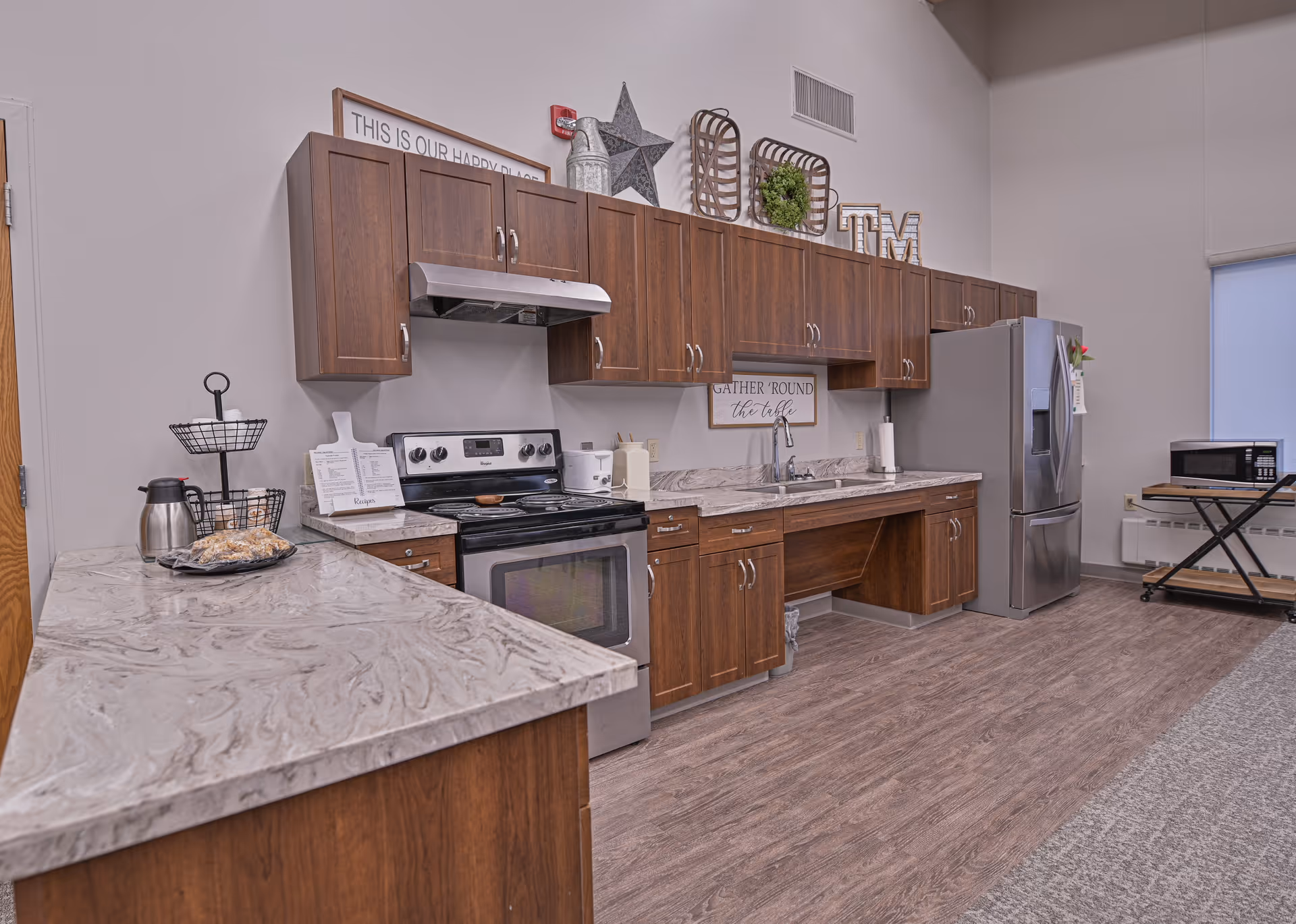 A modern kitchen area in a senior living facility featuring wooden cabinets, a stainless steel oven with a range hood, a stainless steel refrigerator, a microwave on a rolling cart, and a marble-patterned countertop. Decorative items and signs are placed on top of the cabinets and walls, including one that says 'GATHER 'ROUND the table'.