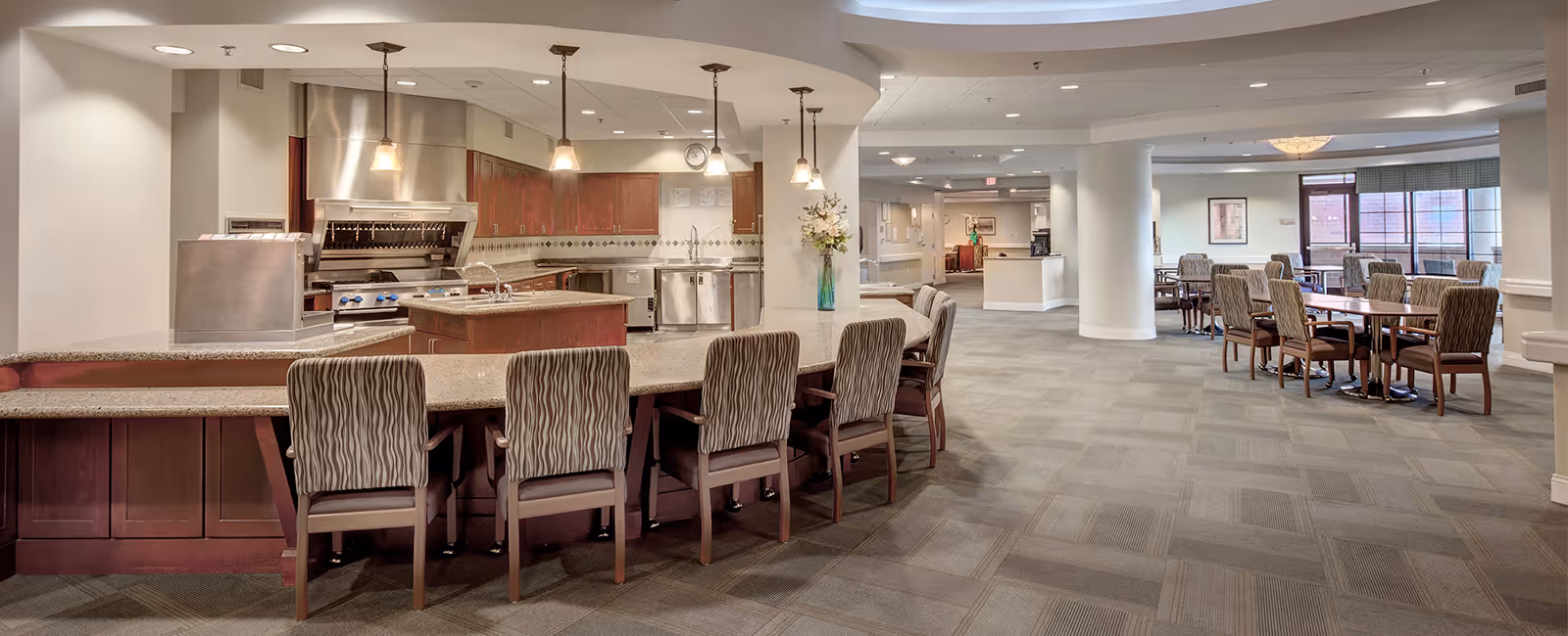 A spacious dining area with multiple tables and chairs, adjacent to an open kitchen featuring stainless steel appliances, wooden cabinets, and a countertop with bar stools. The room has carpeted flooring, white walls, and ceiling lights, creating a bright and welcoming atmosphere.