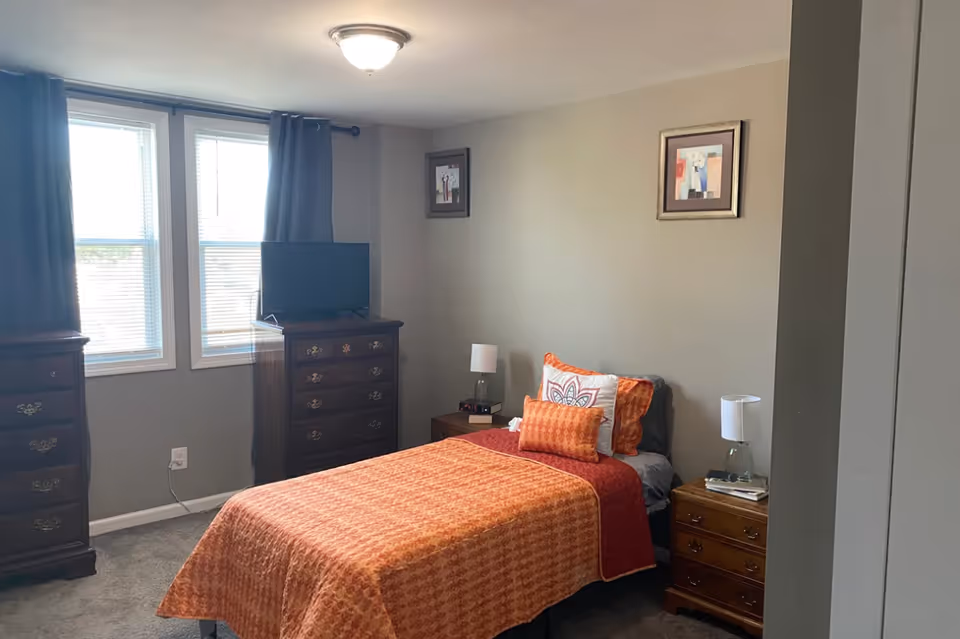 A bedroom with a single bed covered in an orange patterned bedspread and multiple pillows. There are two wooden nightstands on either side of the bed, each with a lamp. A wooden dresser with a TV on top is positioned against the wall near two windows with blinds and dark curtains. The walls are painted beige and have two framed pictures hanging.
