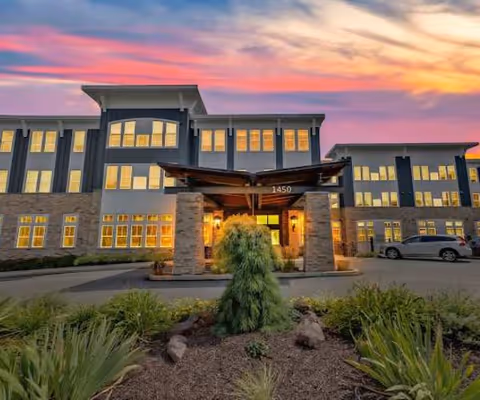 Modern senior living facility exterior with illuminated windows and a covered entrance at sunset