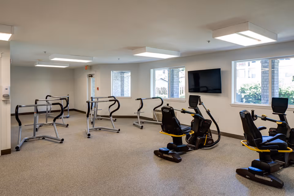 A fitness room in a senior living facility with exercise equipment including stationary recumbent bikes and parallel bars for walking support. The room has large windows letting in natural light and a wall-mounted flat screen TV.