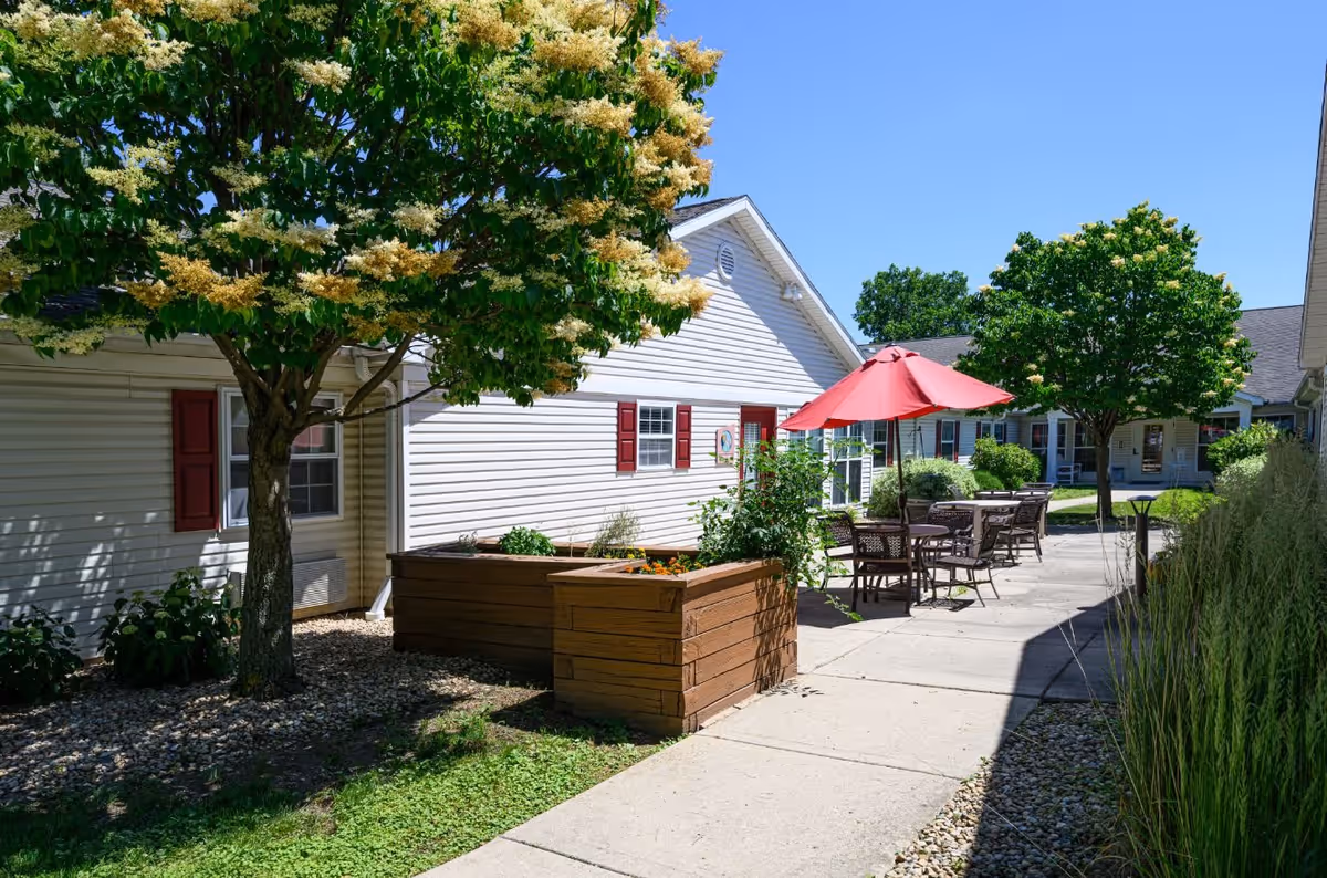Outdoor patio area at Arden Courts - ProMedica Memory Care Community (Glen Ellyn) featuring a paved walkway, wooden planter boxes with plants, metal tables and chairs with a red umbrella, and trees providing shade under a clear blue sky.
