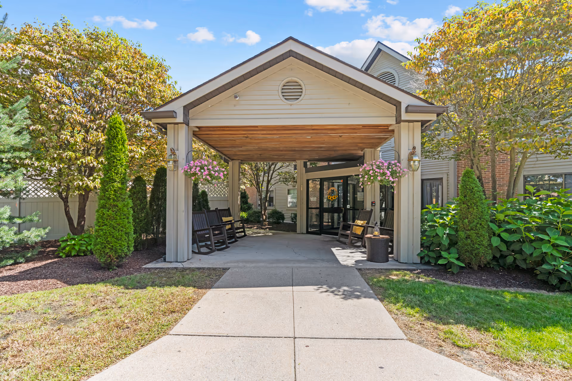 Entrance to a senior living facility with a covered driveway, wooden rocking chairs on either side, hanging flower baskets, and surrounding greenery including trees and shrubs under a clear blue sky.
