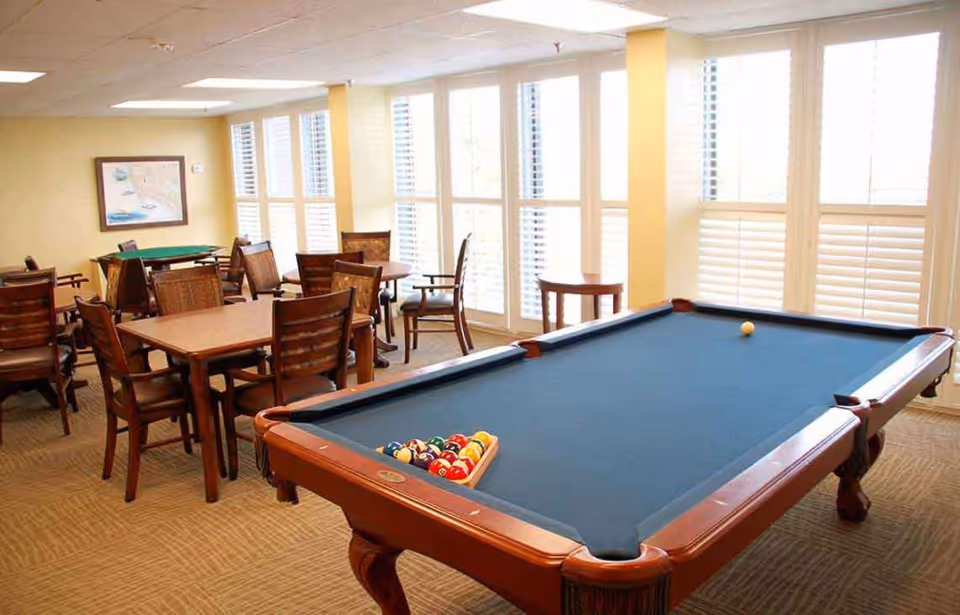 Bright interior game room with a pool table in the foreground and several card tables and chairs by large shuttered windows.