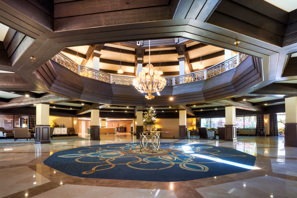 Spacious hotel-style lobby with a large chandelier over a round patterned rug and central floral table beneath a wooden mezzanine balcony.
