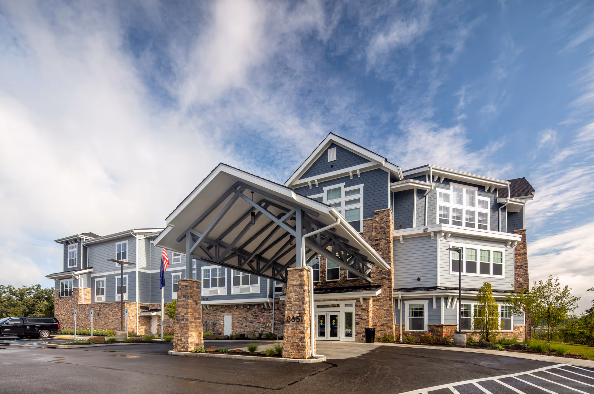 Three-story senior living facility building with a covered porte-cochere entrance, stone accents and blue siding under a partly cloudy sky.