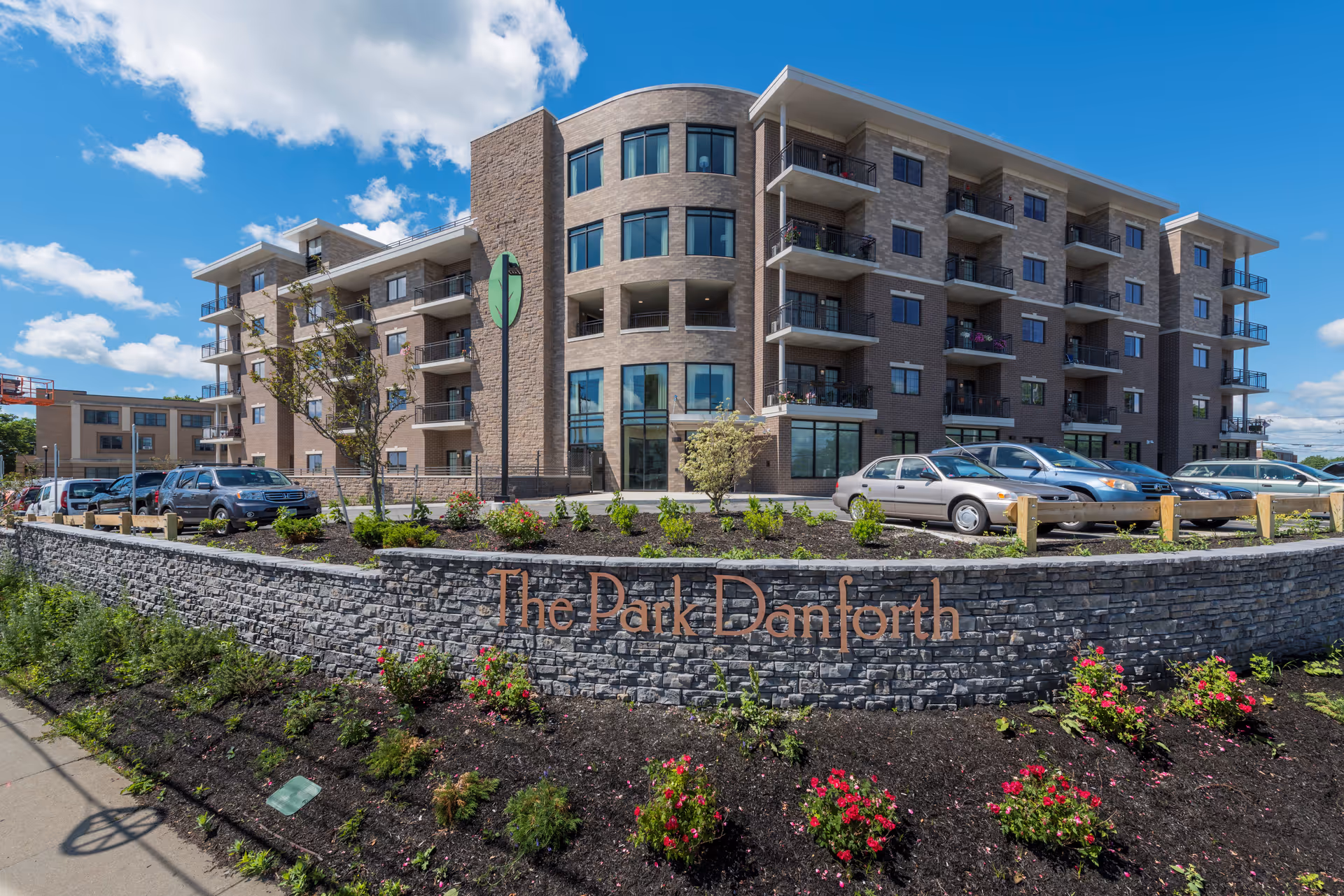 Exterior view of The Park Danforth, a multi-story residential building with balconies, a stone retaining wall with the facility name, landscaped flower beds, and parked cars under a blue sky with scattered clouds.