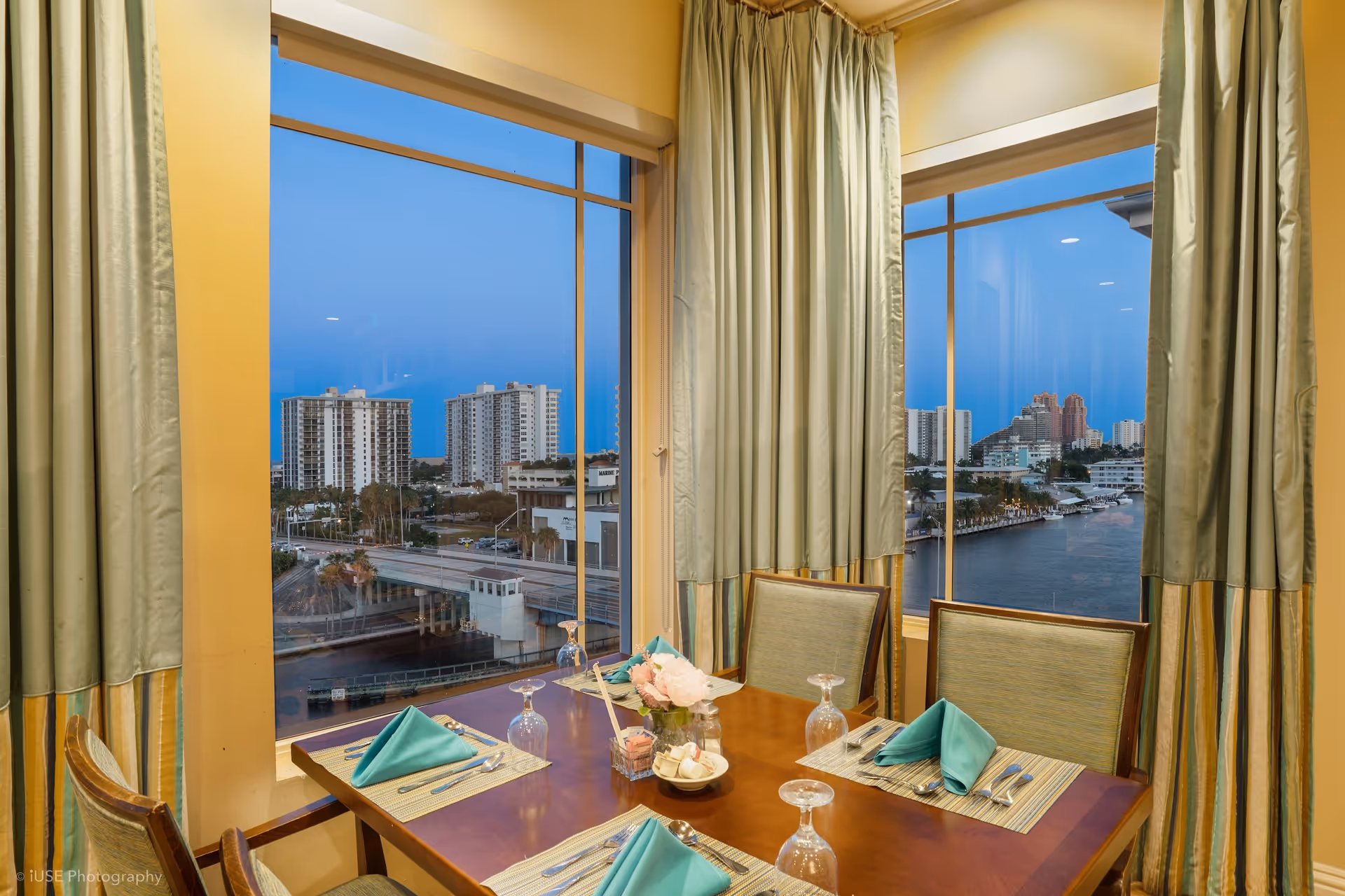 Dining table set for four with teal napkins, glassware, and a small flower centerpiece, positioned near large windows with a view of a cityscape and waterway at dusk. The room has light-colored walls and curtains framing the windows.