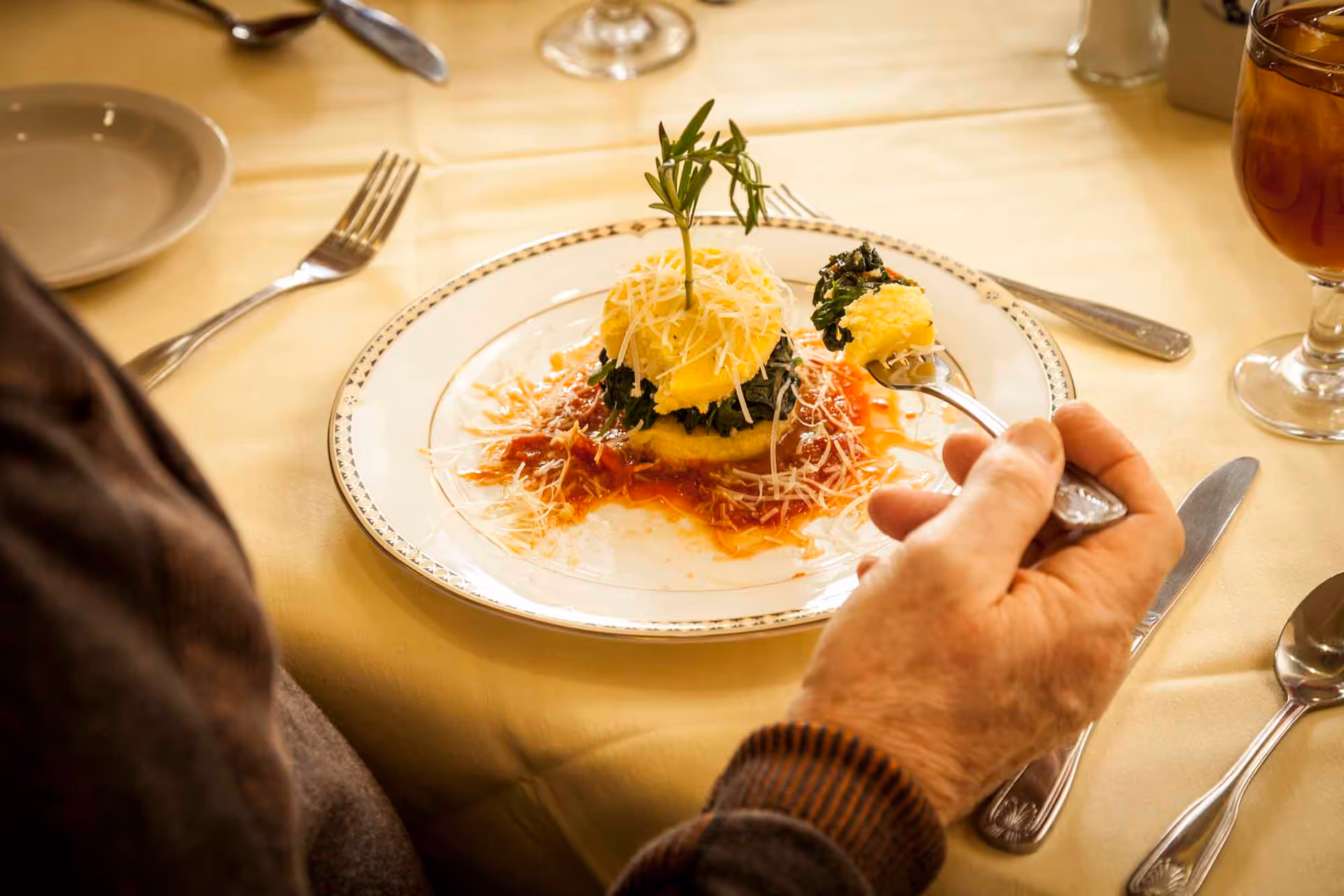 A person holding a fork with a bite of food above a plate containing a gourmet dish garnished with shredded cheese and a sprig of rosemary, set on a yellow tablecloth with silverware and a glass of iced tea nearby.