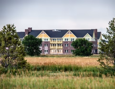 Three-story residential building with yellow and brick facade and balconies seen across a grassy field and trees.