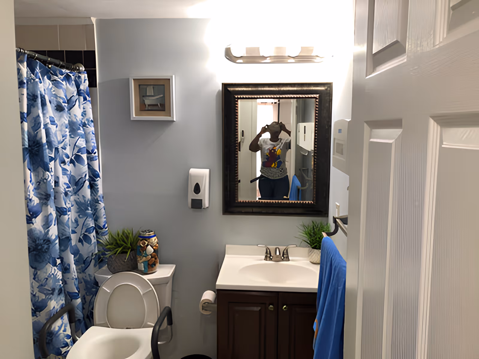 A small bathroom with a white toilet that has a safety frame around it, a sink with a dark wooden cabinet, a mirror above the sink, a blue floral shower curtain, a blue towel hanging on a rack, and some decorative plants and items on the toilet tank. A person is visible in the mirror taking the photo.