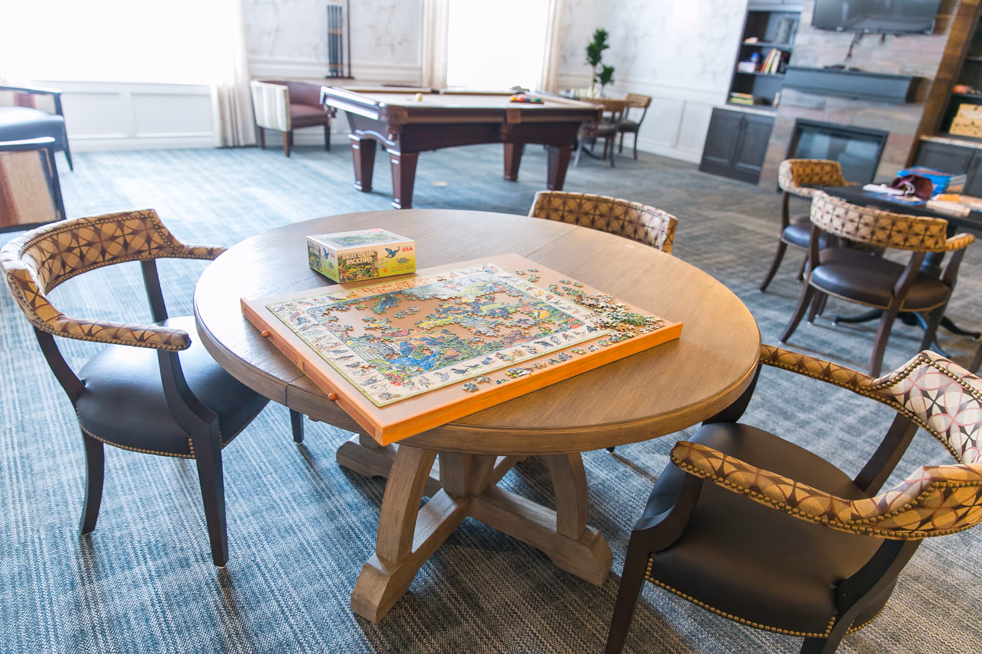 A round wooden table with a partially completed jigsaw puzzle and its box on top, surrounded by four cushioned chairs with patterned backs. In the background, there is a pool table, additional seating, a fireplace, and a television mounted above a dark wooden cabinet in a spacious, carpeted room with large windows.