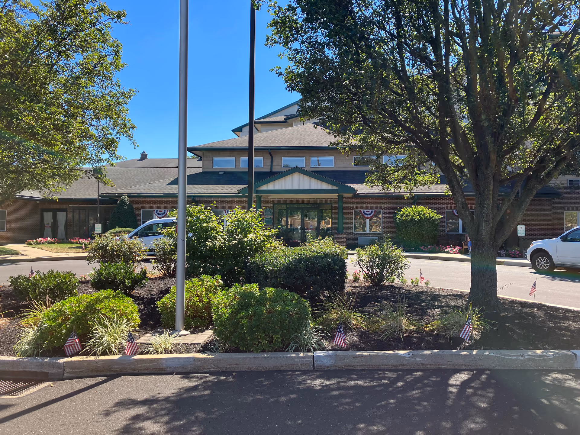 Front exterior view of Gloria Dei Estates facility with a landscaped garden area featuring bushes, small plants, and American flags. The building has a brick facade with a green roof and multiple windows. There are two flagpoles in front of the garden and a tree on the right side. A white vehicle is parked on the right side of the building.