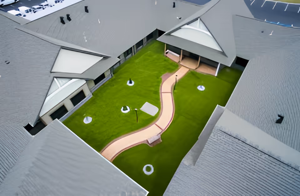 Aerial view of a green courtyard with a winding paved pathway, surrounded by the gray rooftops of a building. The courtyard features small circular garden beds and a covered seating area.