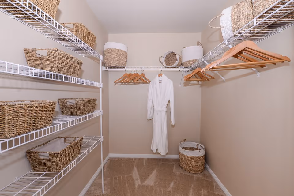 A walk-in closet with beige walls and carpeted floor. The closet has white wire shelving on the left and right walls, holding several woven baskets. Wooden hangers are hanging on the right side and the back wall, with a white bathrobe hanging in the center. Additional woven baskets are placed on the top shelves and the floor.