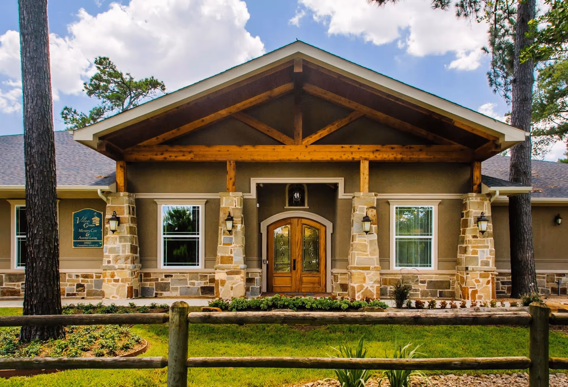 Front exterior view of Village Green Memory Care Community building with stone pillars, wooden double doors, windows on either side, and a wooden fence in the foreground under a partly cloudy sky.