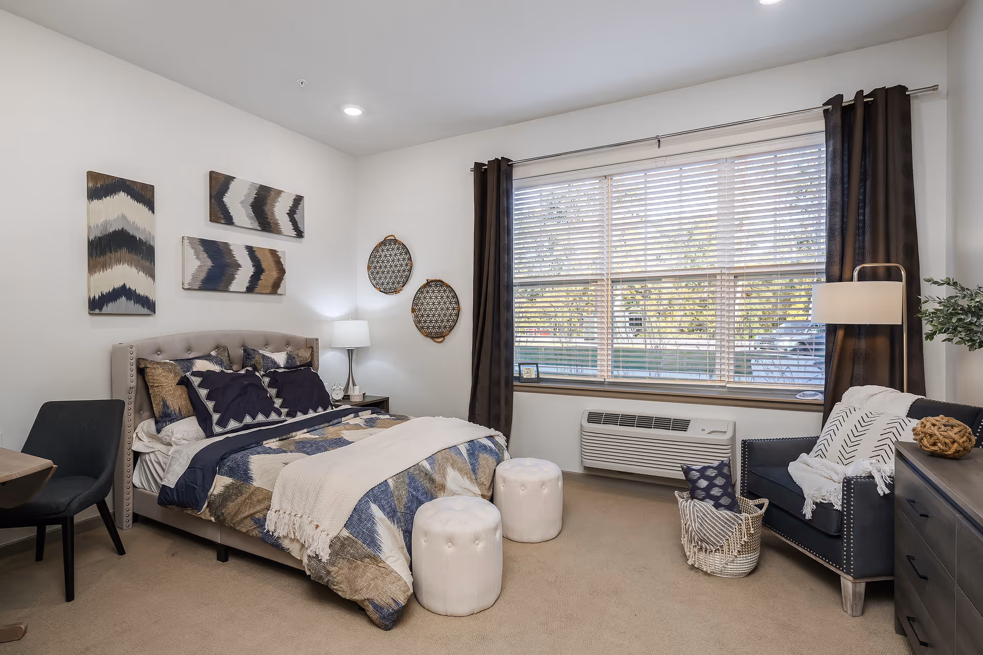 A cozy bedroom with a large window covered by blinds and dark curtains. The room features a bed with patterned bedding and multiple pillows, two white tufted ottomans at the foot of the bed, a dark upholstered armchair with a white throw and pillows, a basket with a blanket and pillow, a nightstand with a lamp, and decorative wall art above the bed and on the adjacent wall.