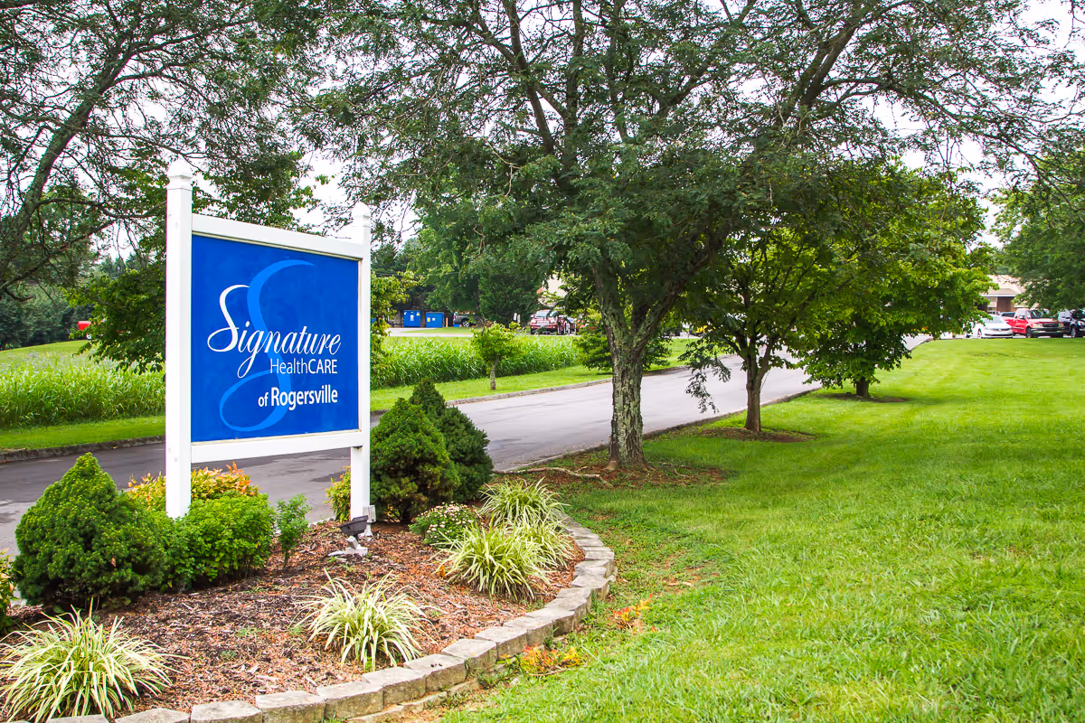 A landscaped lawn and driveway with a blue 'Signature HealthCARE of Rogersville' sign in front of trees and parked cars.