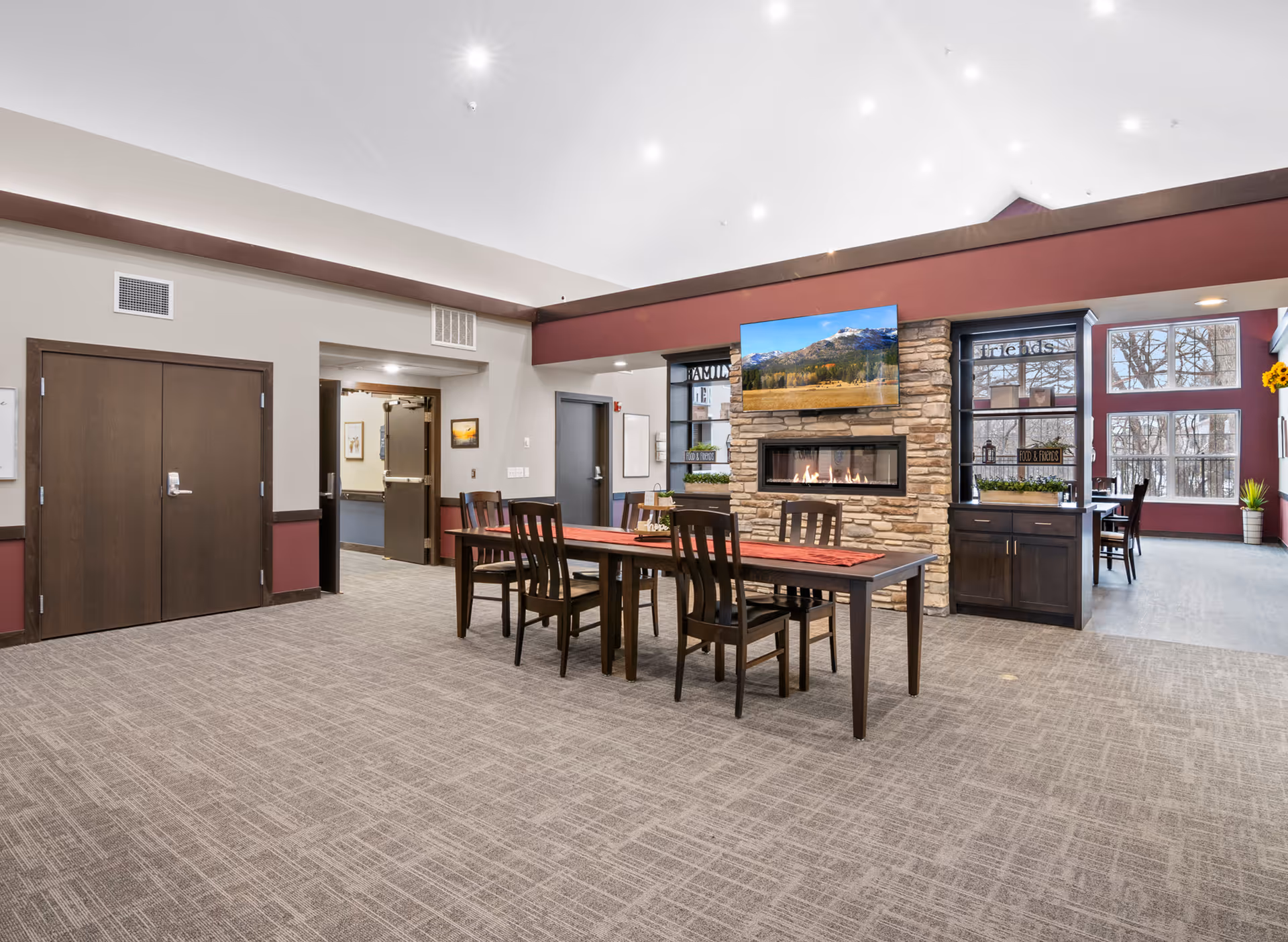 A spacious common area in a senior living facility featuring a long wooden table with six chairs, a stone fireplace with a flat-screen TV mounted above it, and large windows letting in natural light. The room has a high ceiling with recessed lighting and a carpeted floor. There are decorative plants and signs on shelves near the fireplace.