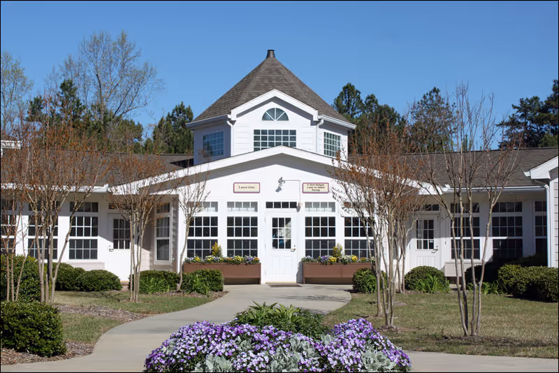 Front entrance of a white senior living building with a central cupola, walkway, and purple flowers in the foreground.
