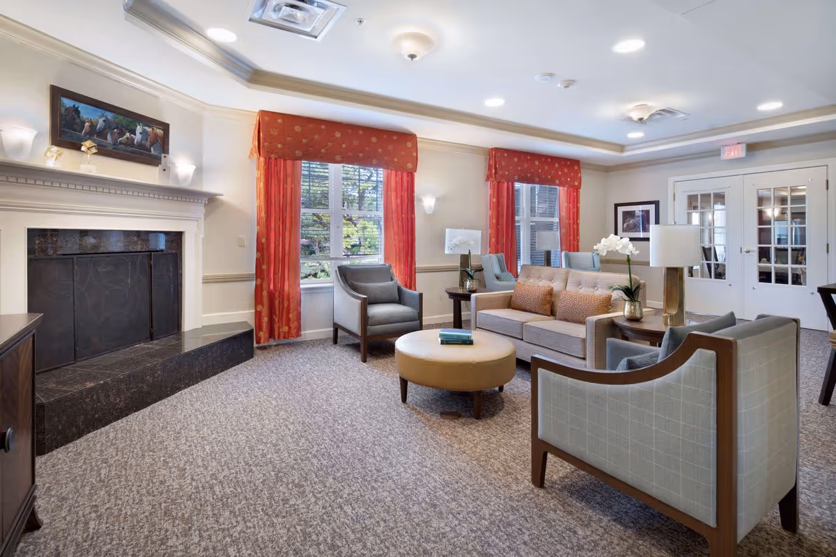 A bright senior living facility lounge with sofas, armchairs, a round ottoman, a fireplace, and red window curtains.