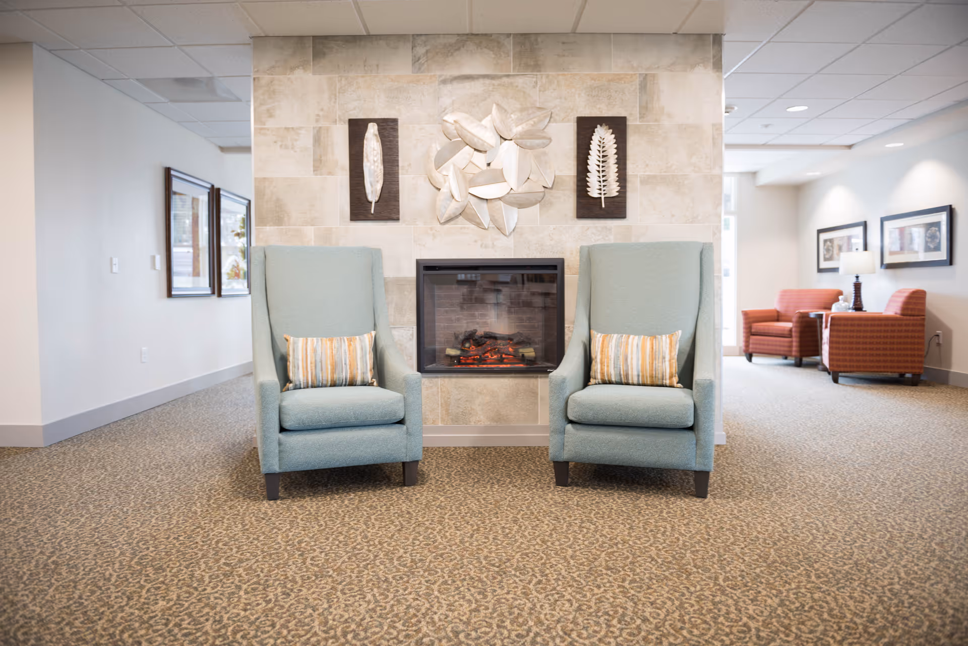 Two teal armchairs flanking a built-in electric fireplace with decorative wall art in a bright senior living lounge area.