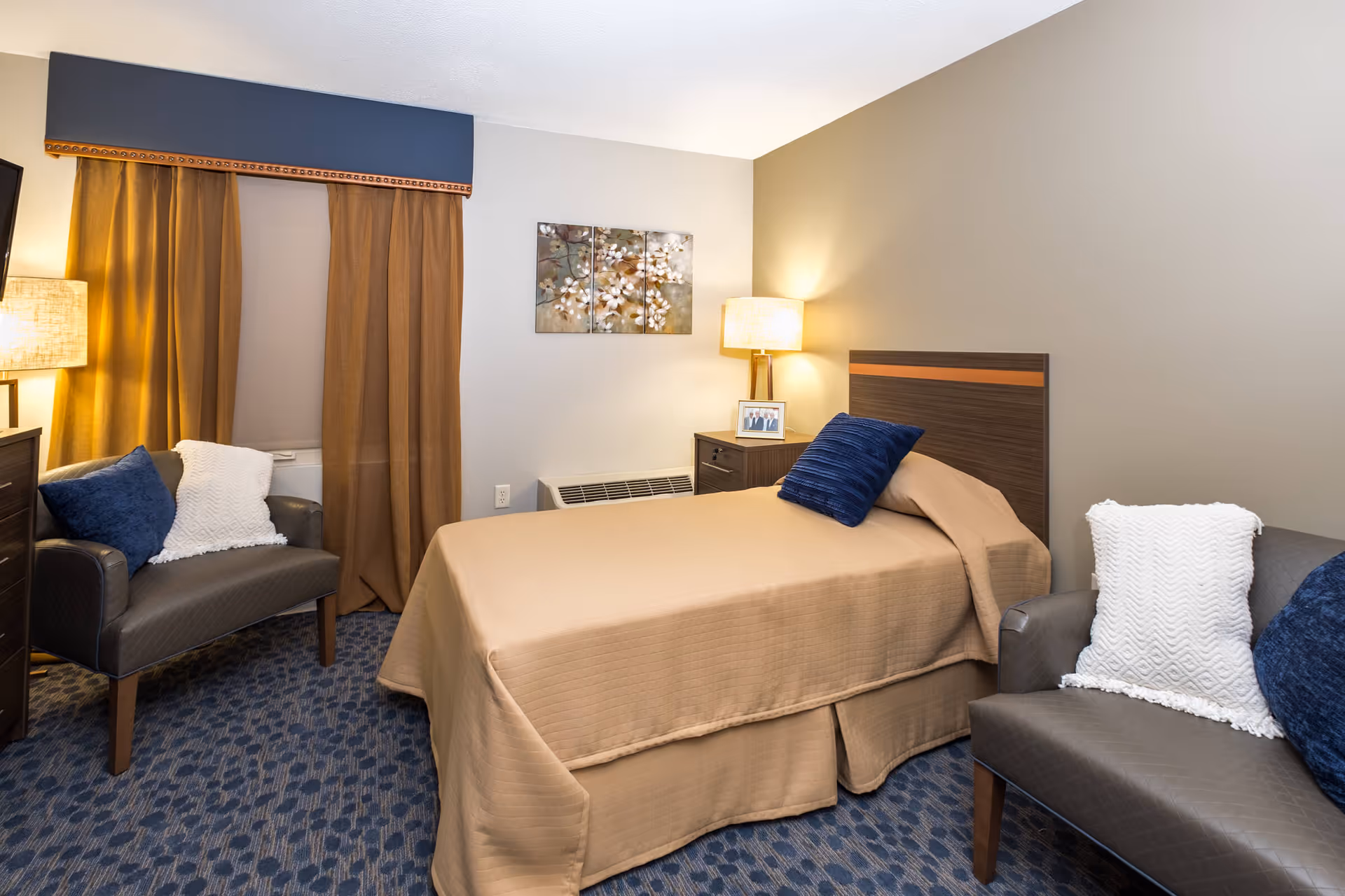 Well-lit single-occupancy bedroom with a bed, two armchairs, a nightstand with a lamp, and a curtained window.